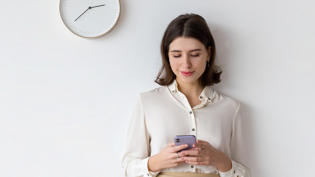 Vrouw bezig met telefoon naast een klok aan de muur.